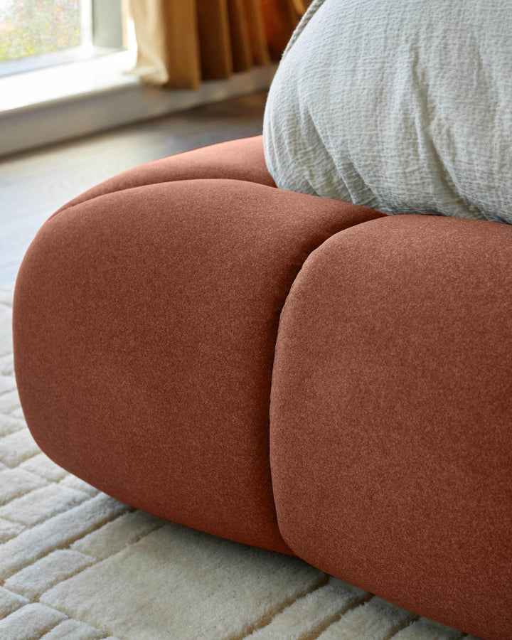 Toffee-red ottoman bed with rounded, cushioned sides, shown from close-up angle in bedroom setting.