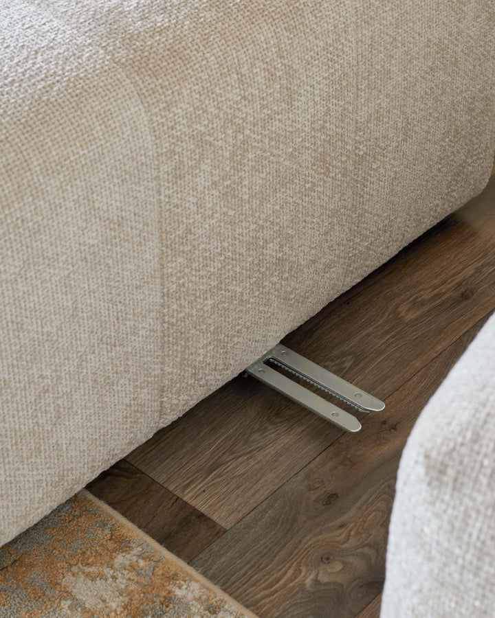 Close-up of a neutral-toned sofa with a metallic reclining mechanism visible against a wood floor and patterned rug.