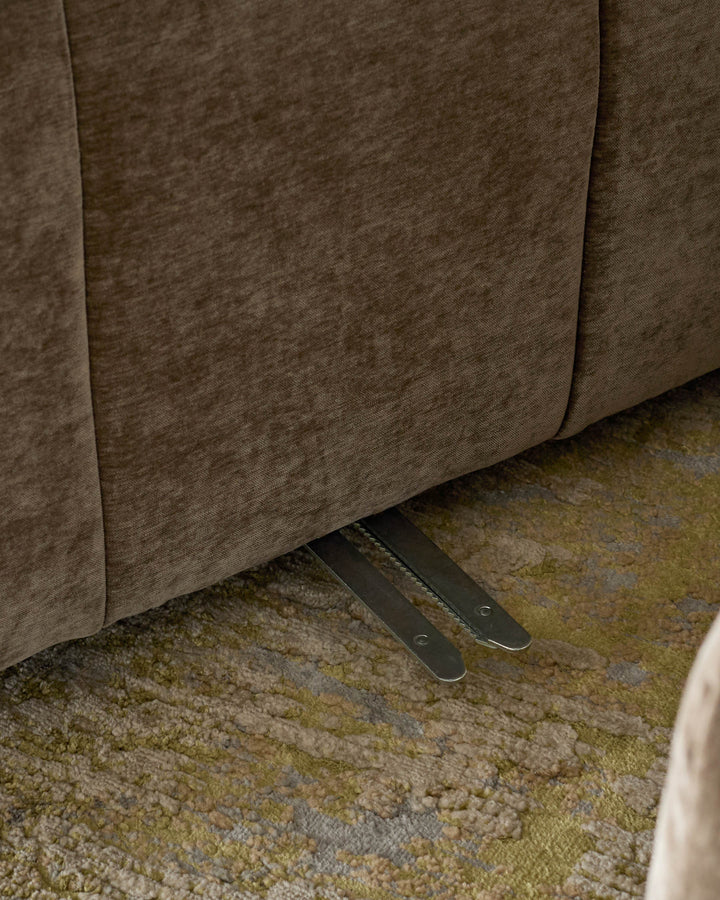 Close-up of a brown fabric sofa, with metal reclining mechanism visible underneath. Displayed on a textured beige & green rug.