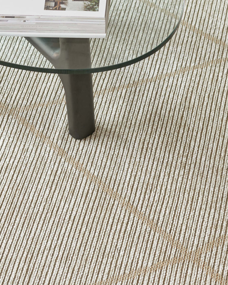 Modern round glass-top coffee table with a single cylindrical metal leg on a textured beige and white striped area rug.