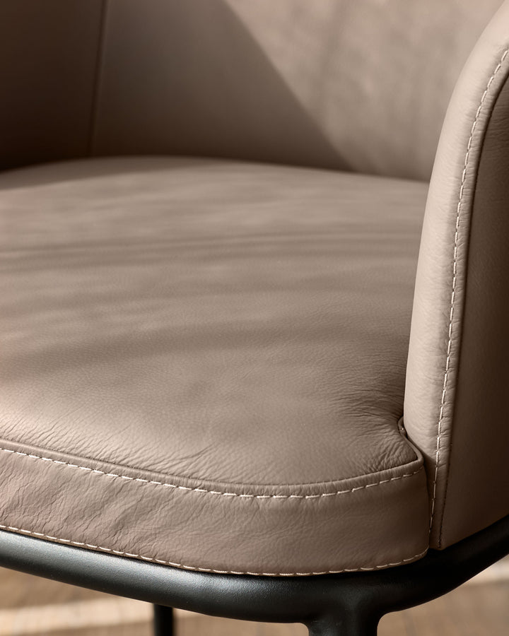 Logan dining chair in mocha leather, close-up of seat and backrest showing stitched seams and curved armrest detail.