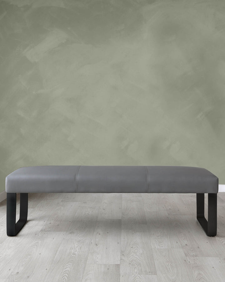 Modern minimalist grey upholstered bench with black legs, set on a light wooden floor against a textured green-grey wall.
