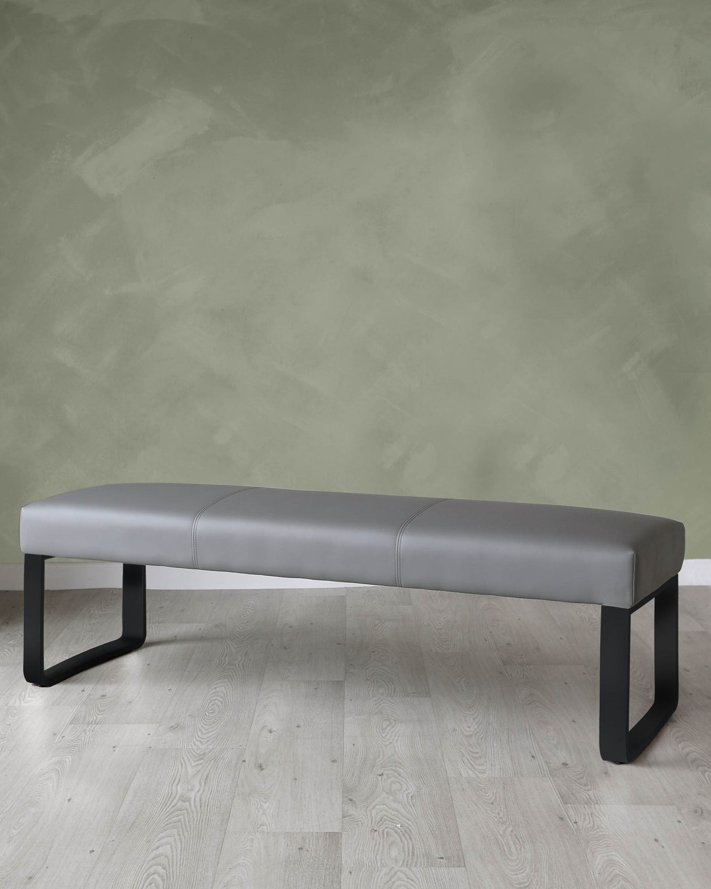 Modern grey upholstered bench with black metal legs on a light wood floor against a textured green wall.