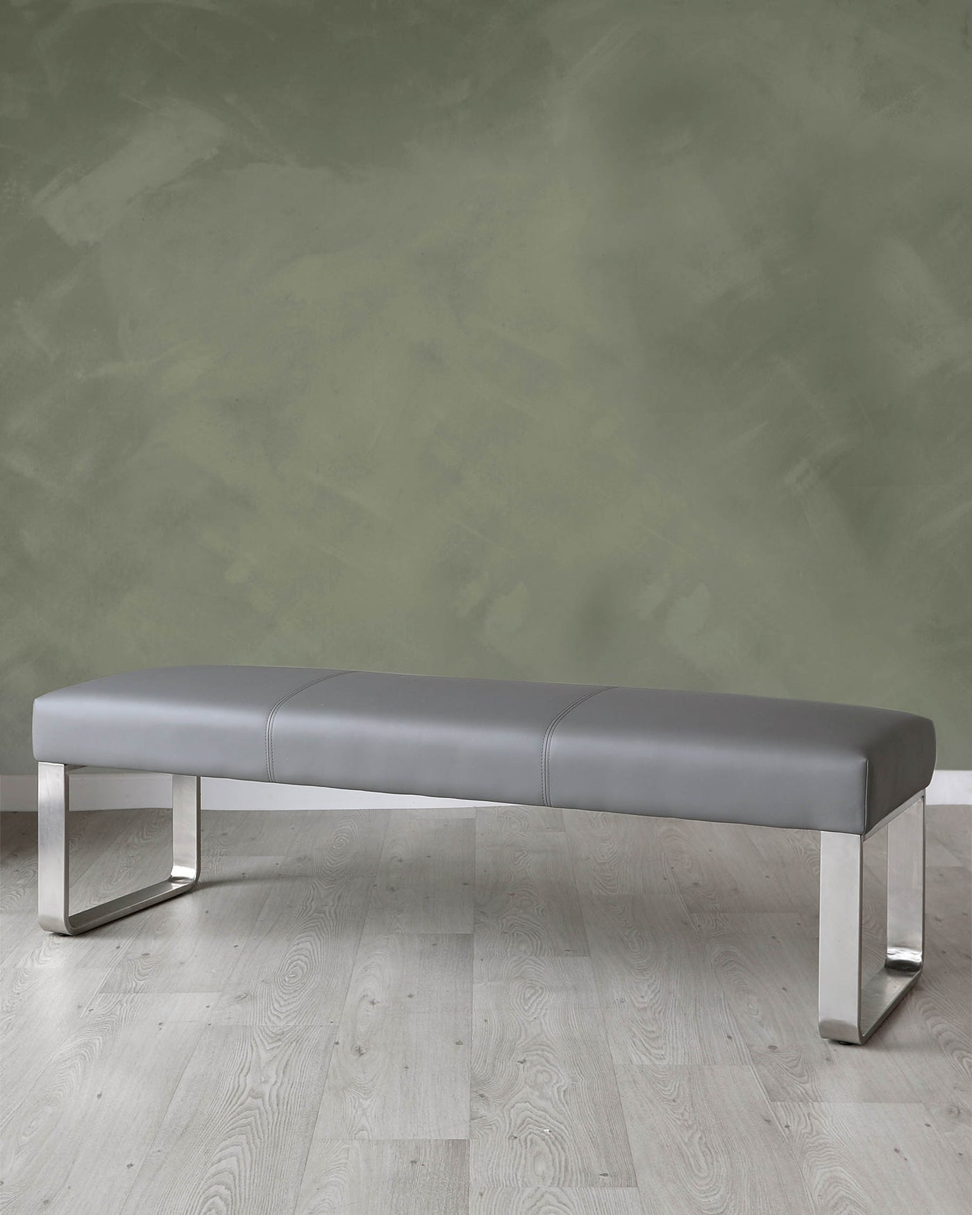 Modern grey leather bench with sleek chrome legs on a light wooden floor against a textured green wall.
