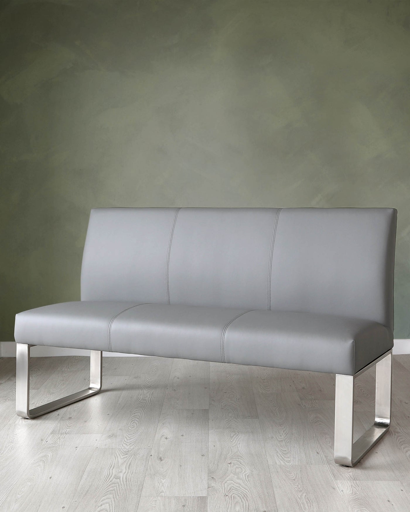 A modern grey leather bench with a minimalist design featuring a straight backrest, seat with subtle stitching detail, and sleek chrome U-shaped legs positioned on a light wood floor against a textured olive green wall.