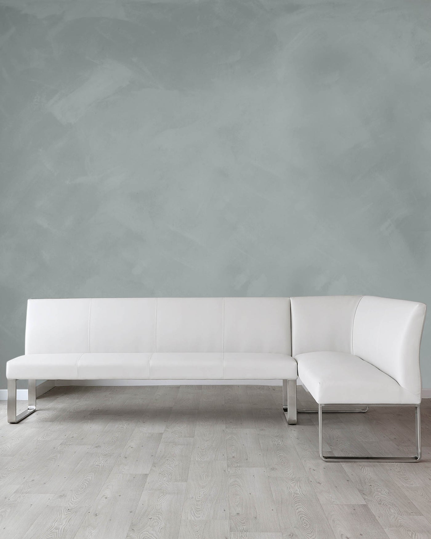 Modern white leather sectional sofa with a minimalist design, featuring clean lines, cushioned seating, and sleek metal legs, set against a textured grey wall and light wooden flooring.