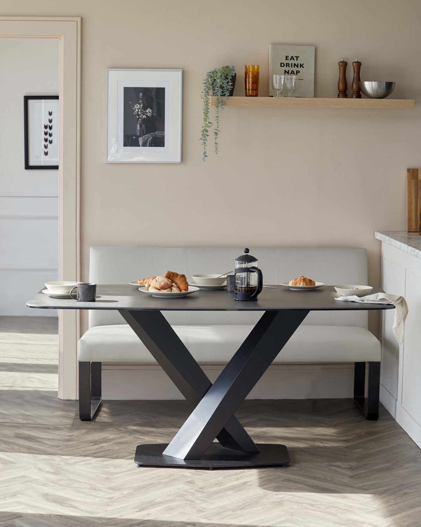 Modern minimalist dining set featuring a rectangular table with a sleek dark stone or wood finish top and a bold, black metallic X-shaped base, accompanied by a matching long bench with light beige upholstery.
