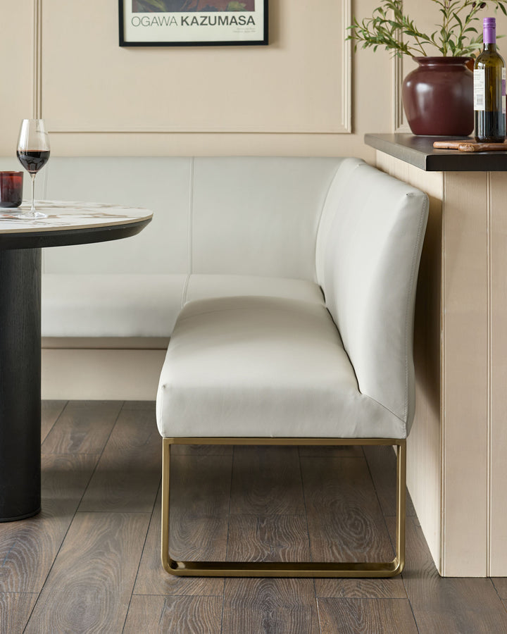 White faux leather dining chair with brushed brass legs, shown in a modern dining setting next to a round table.
