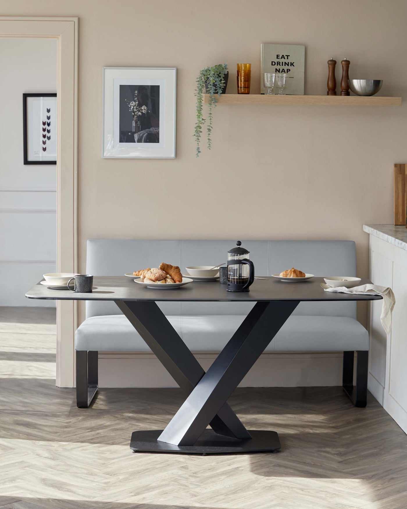 Modern dining room furniture featuring a dark-toned tabletop with a striking X-shaped metal base paired with a long, contemporary bench with grey upholstery. A wooden floating shelf is mounted above, displaying decorative items.