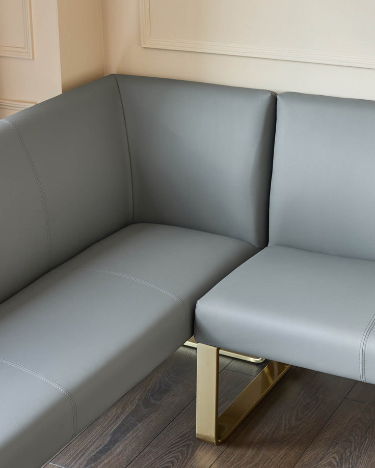 Grey faux leather corner dining bench with brushed brass legs, shown at an angle in a bright room with beige walls.