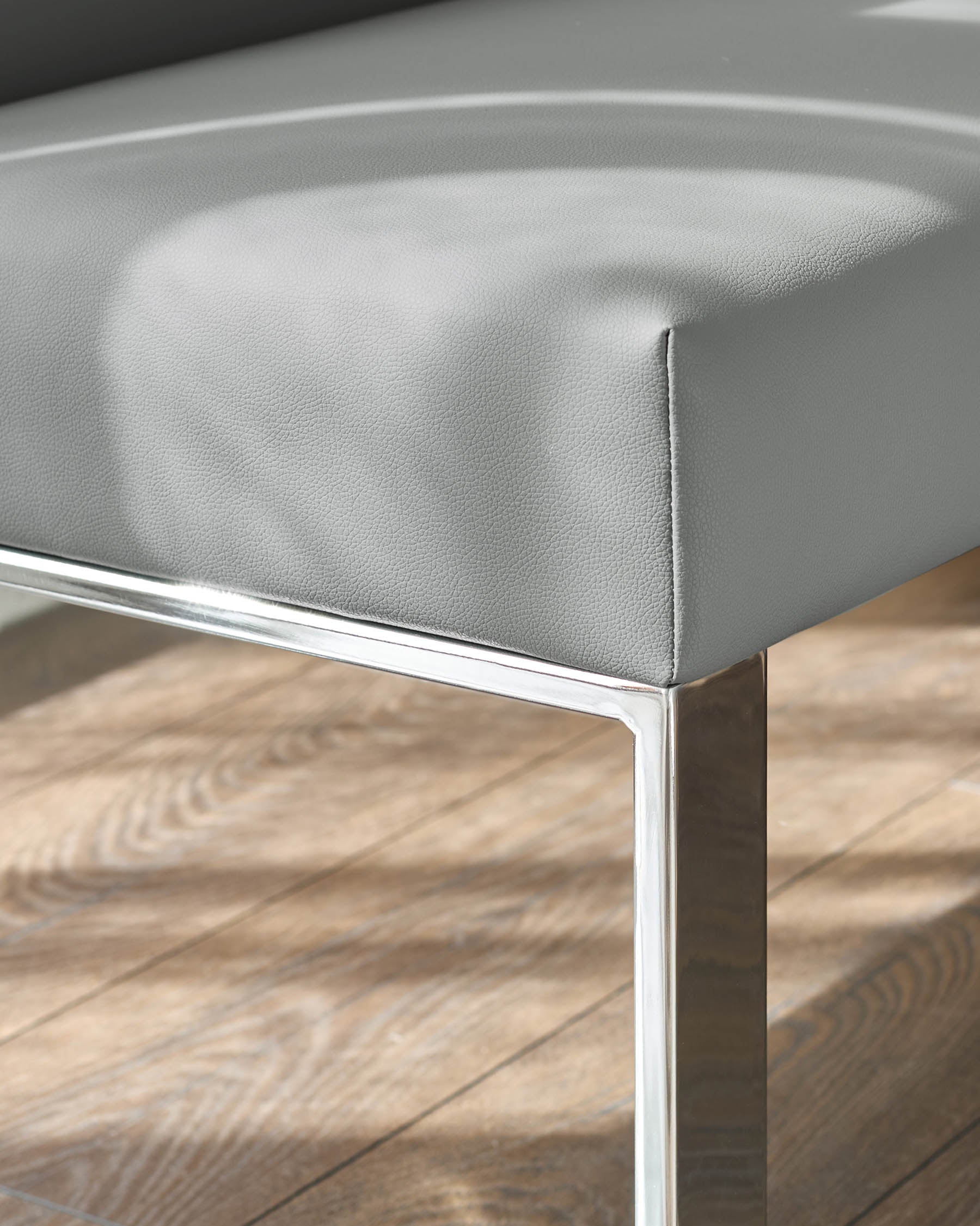 Grey faux leather dining bench with chrome legs, close-up detail view showing textured upholstery and metal frame on wooden floor.