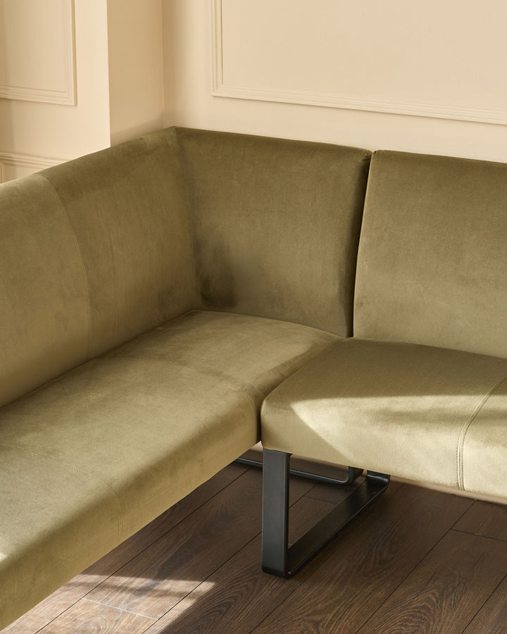 Olive green velvet corner bench with black metal legs, right-hand configuration, positioned in a bright corner space.