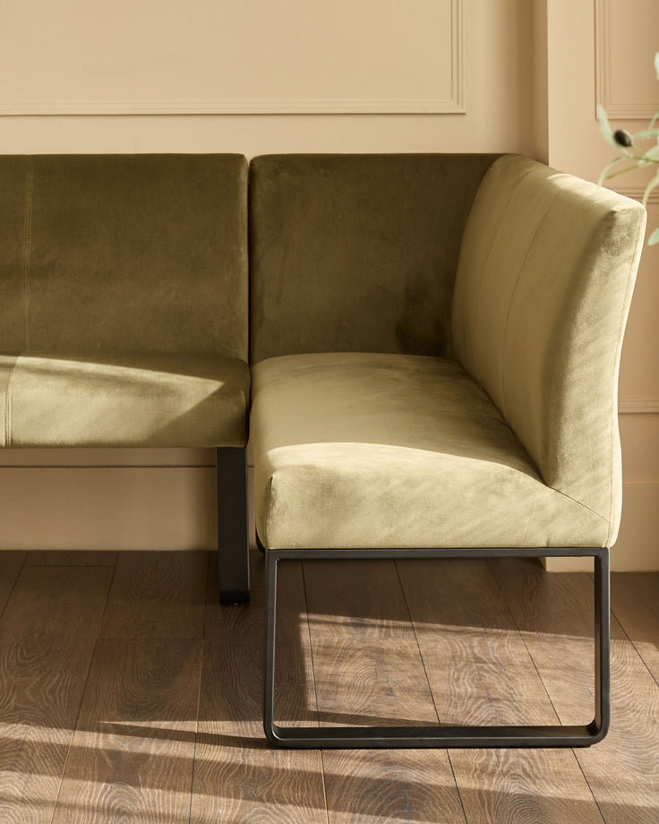 Olive green velvet corner bench with black metal legs, shown from side angle in bright sunlight on wooden floor.