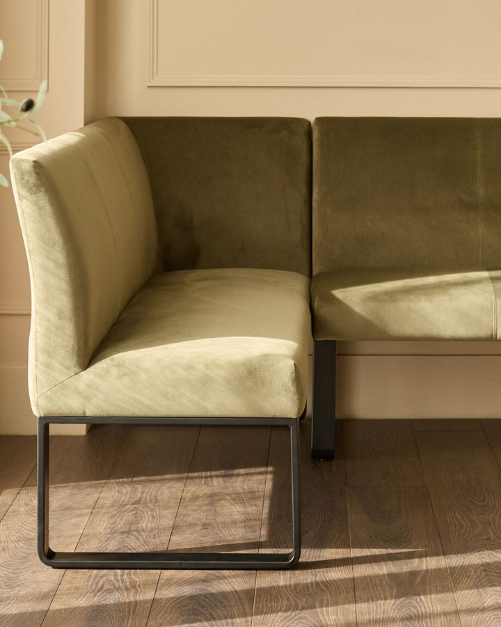 Olive green velvet corner bench with black metal legs, shown at an angle in a bright, neutral interior space.