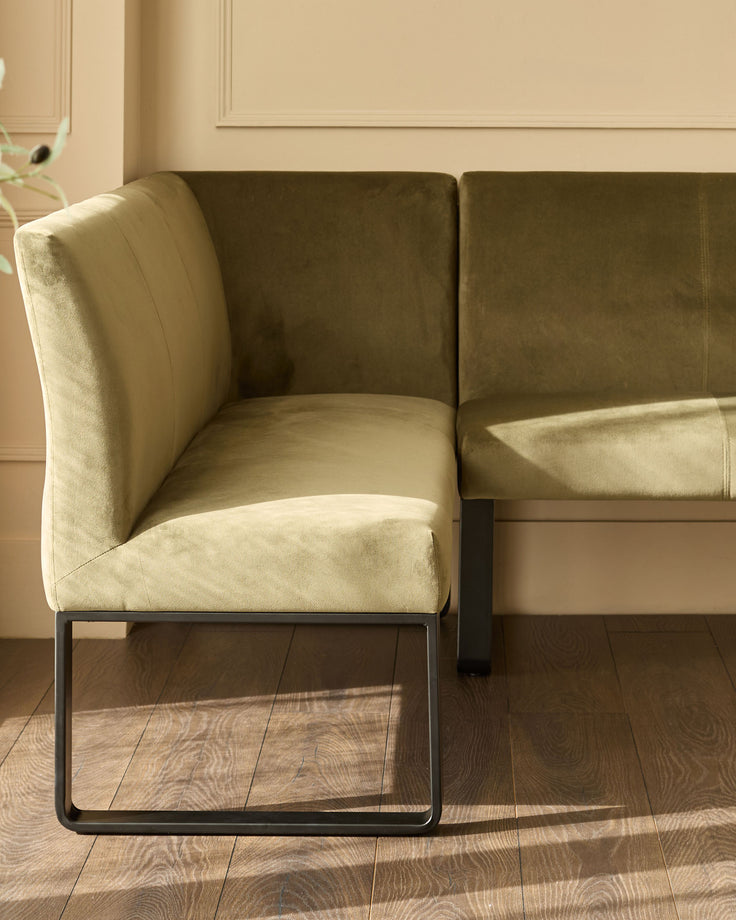 Olive green velvet corner bench with black metal legs, shown at an angle in a bright, neutral interior space.