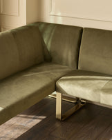 Olive green corner bench with brushed brass legs, angled view showing curved backrest and seating in minimalist interior.