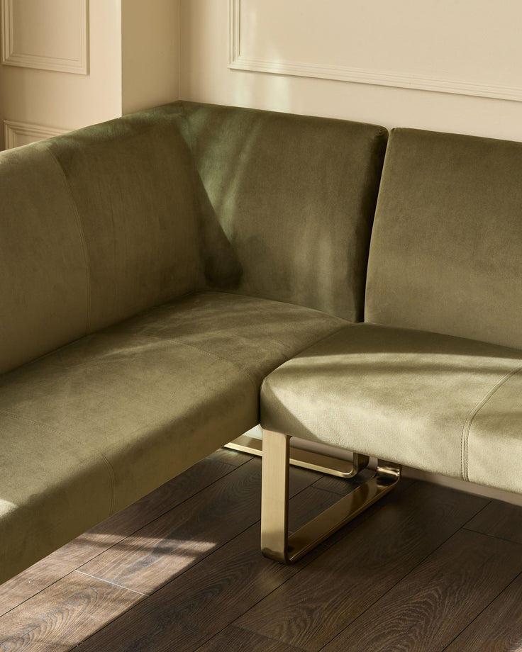 Olive green corner bench with brushed brass legs, angled view showing curved backrest and seating in minimalist interior.