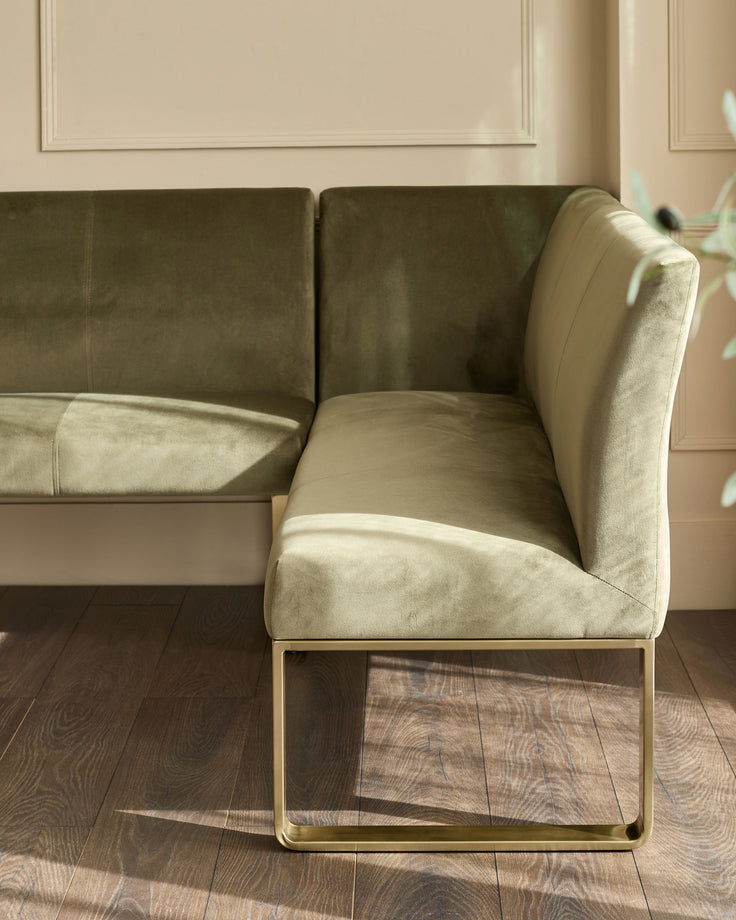 Olive green upholstered corner bench with brushed brass legs, shown at an angle in a neutral room setting.
