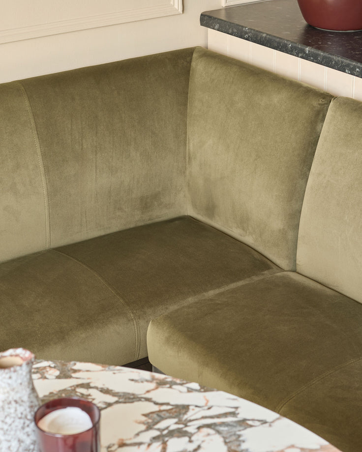 Olive green upholstered corner bench with curved armrests and stainless steel leg, shown in living room setting.