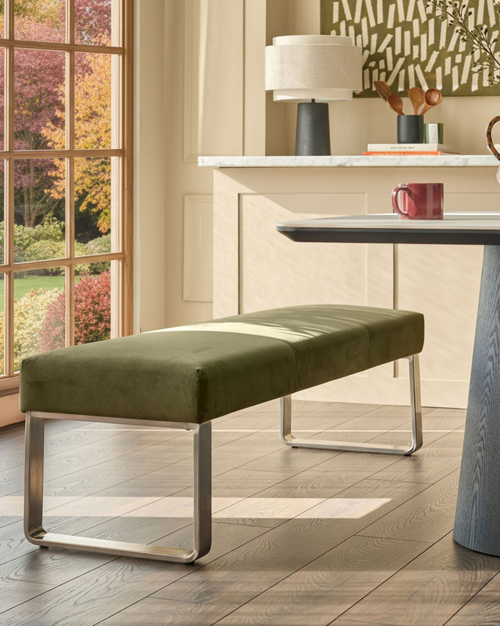 Olive green velvet bench with stainless steel loop frame, positioned in a bright room with autumn views through windows.