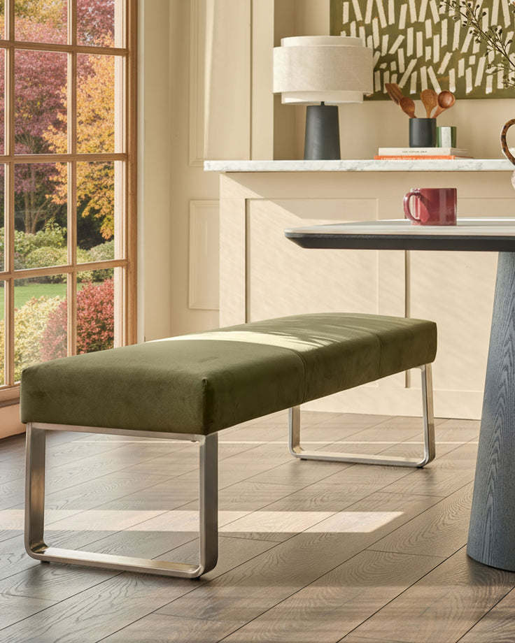 Olive green velvet bench with stainless steel loop frame, positioned in a bright room with autumn views through windows.