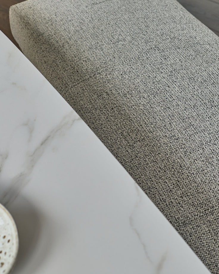 Grey fabric 3-seater bench without backrest, shown at an angle on white marble floor.