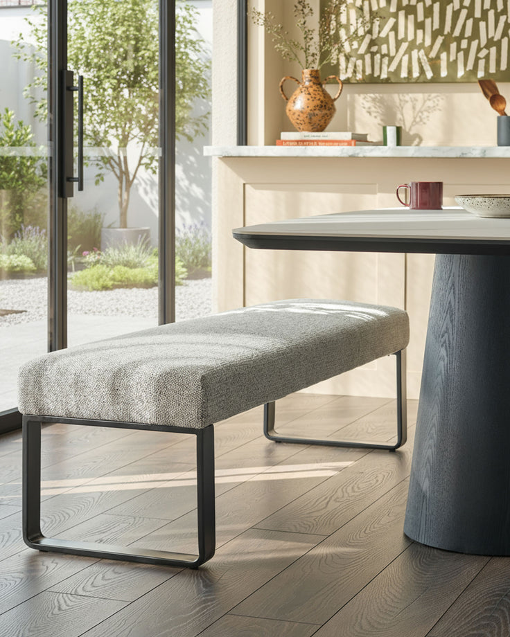 Grey fabric bench with black metal frame, positioned in a modern interior space with wooden flooring and shelving.