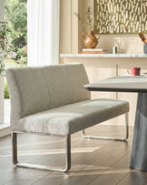 Grey fabric bench with backrest and stainless steel frame, positioned at dining table in bright, modern interior.