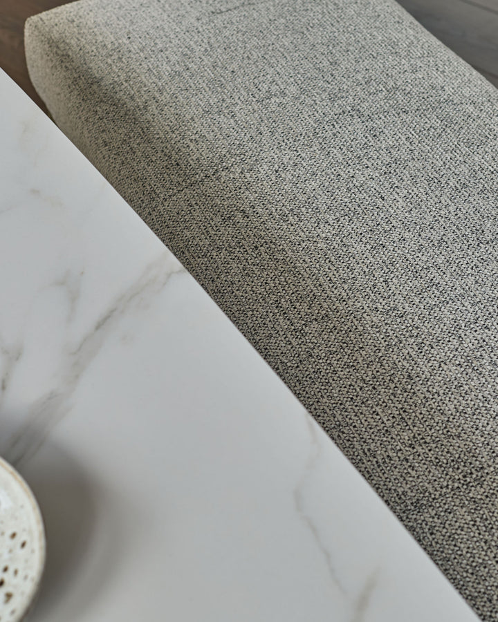Grey fabric 3-seater bench without backrest, shown at an angle displaying the textured upholstery and brushed brass base.