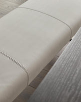 Close-up of light grey upholstered bench seat with stitched seam detail and black legs on wooden floor.