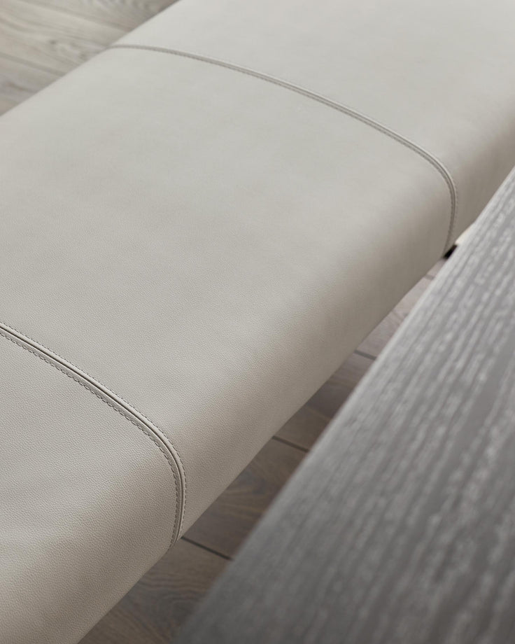 Close-up of light grey upholstered bench seat with stitched seam detail and black legs on wooden floor.