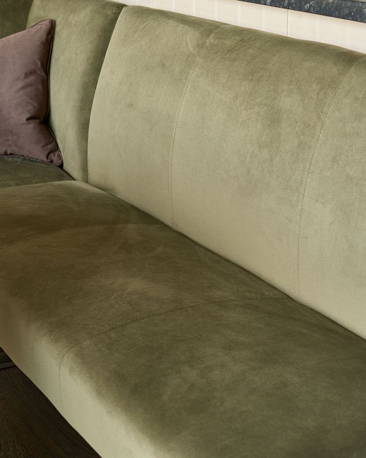 Olive green velvet corner sofa with cushioned backrest and seat, shown from a side angle with brown accent pillow.