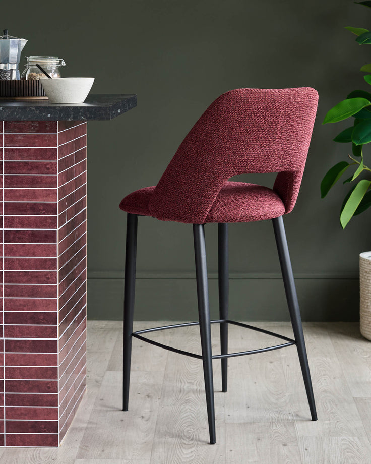 A red fabric bar stool with a black metal frame is pictured against a dark green wall and wooden flooring.