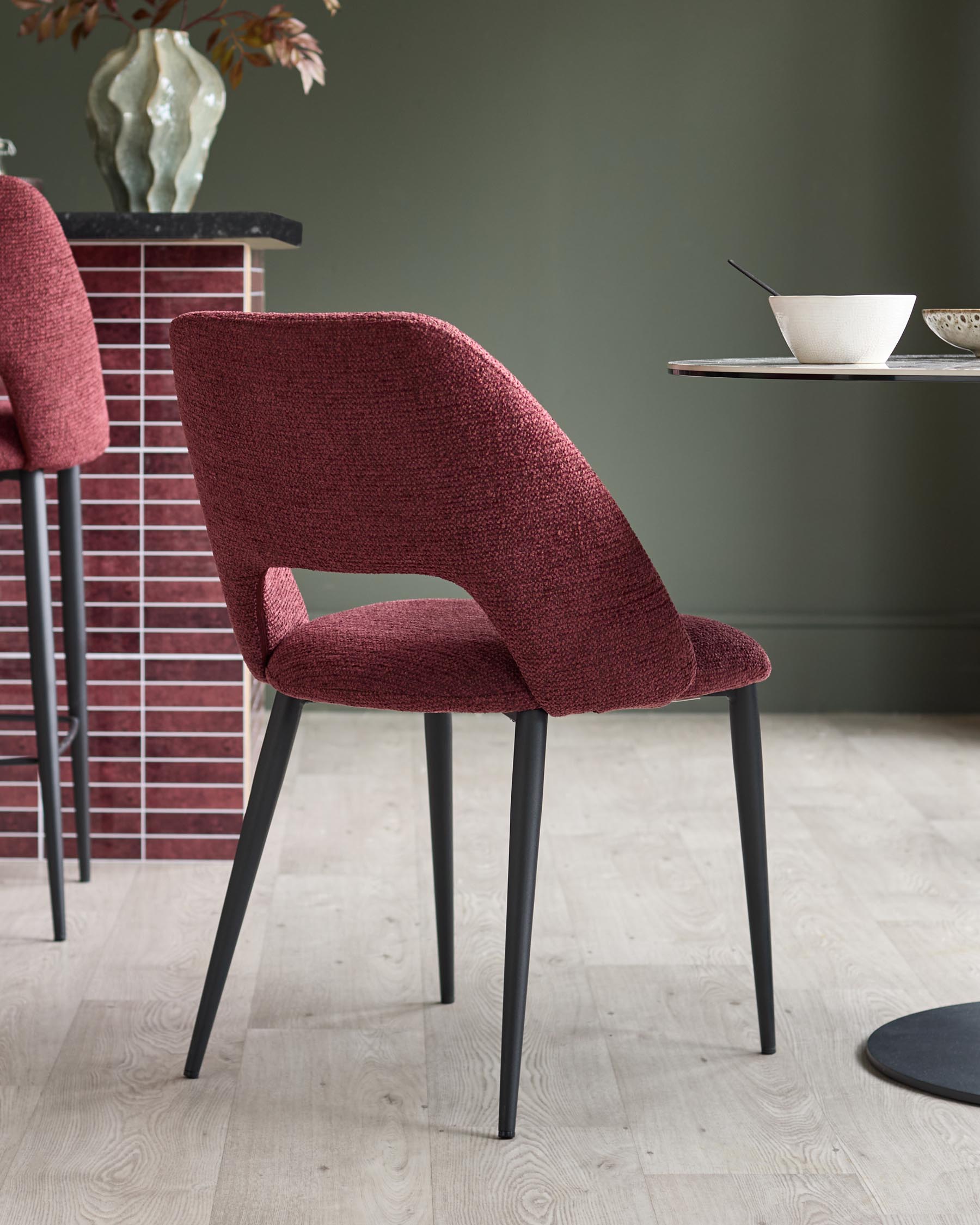 A modern dining chair in red upholstery, with black metal legs, alongside a table and counter with tiled backsplash.