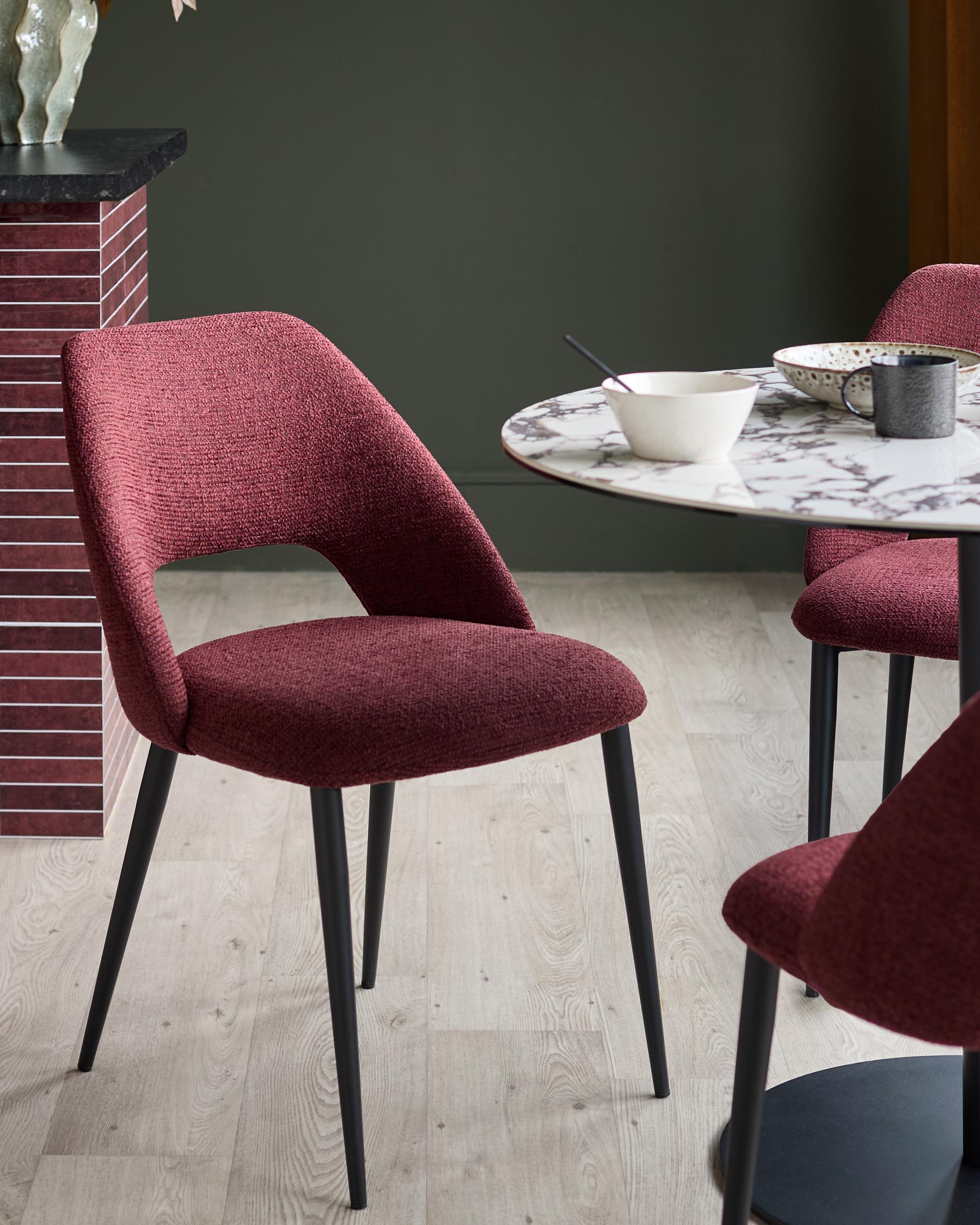 Stylish dining setting with modern chairs upholstered in a textured, deep red fabric and sleek black legs, around a marble-effect table.