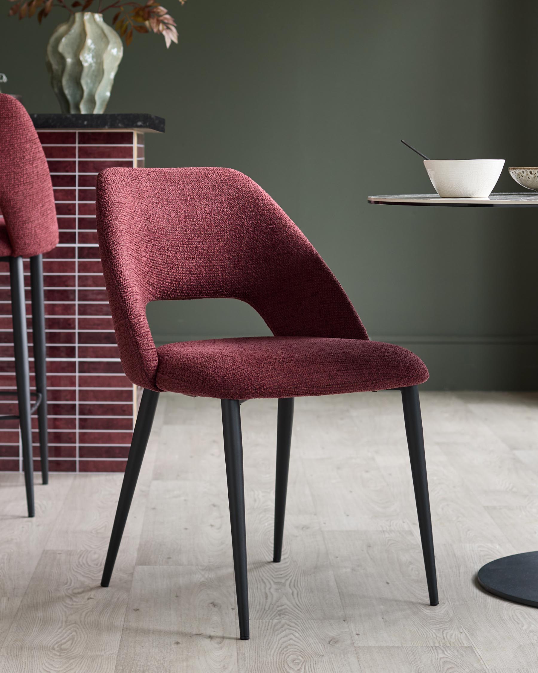 A stylish, modern chair upholstered in red fabric with sleek black legs, next to a matching bar stool and round table.