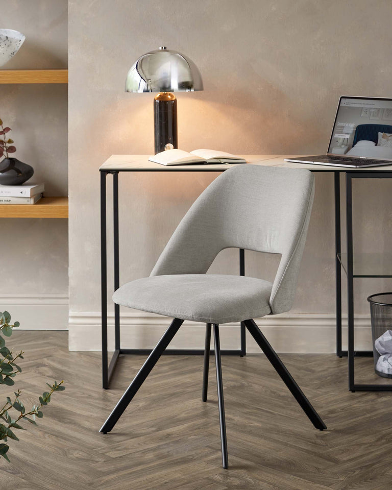 Contemporary light grey upholstered chair with a curved backrest and angled black metal legs, paired with a sleek black metal frame desk featuring a light wooden top. A modern silver dome table lamp is positioned on the desk, illuminating the workspace.