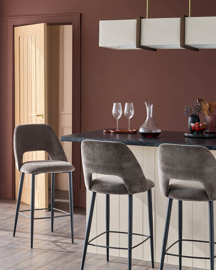 Three brown velvet bar stools sit at a kitchen island with wine glasses and decanter on a dark worktop, under a long pendant light.