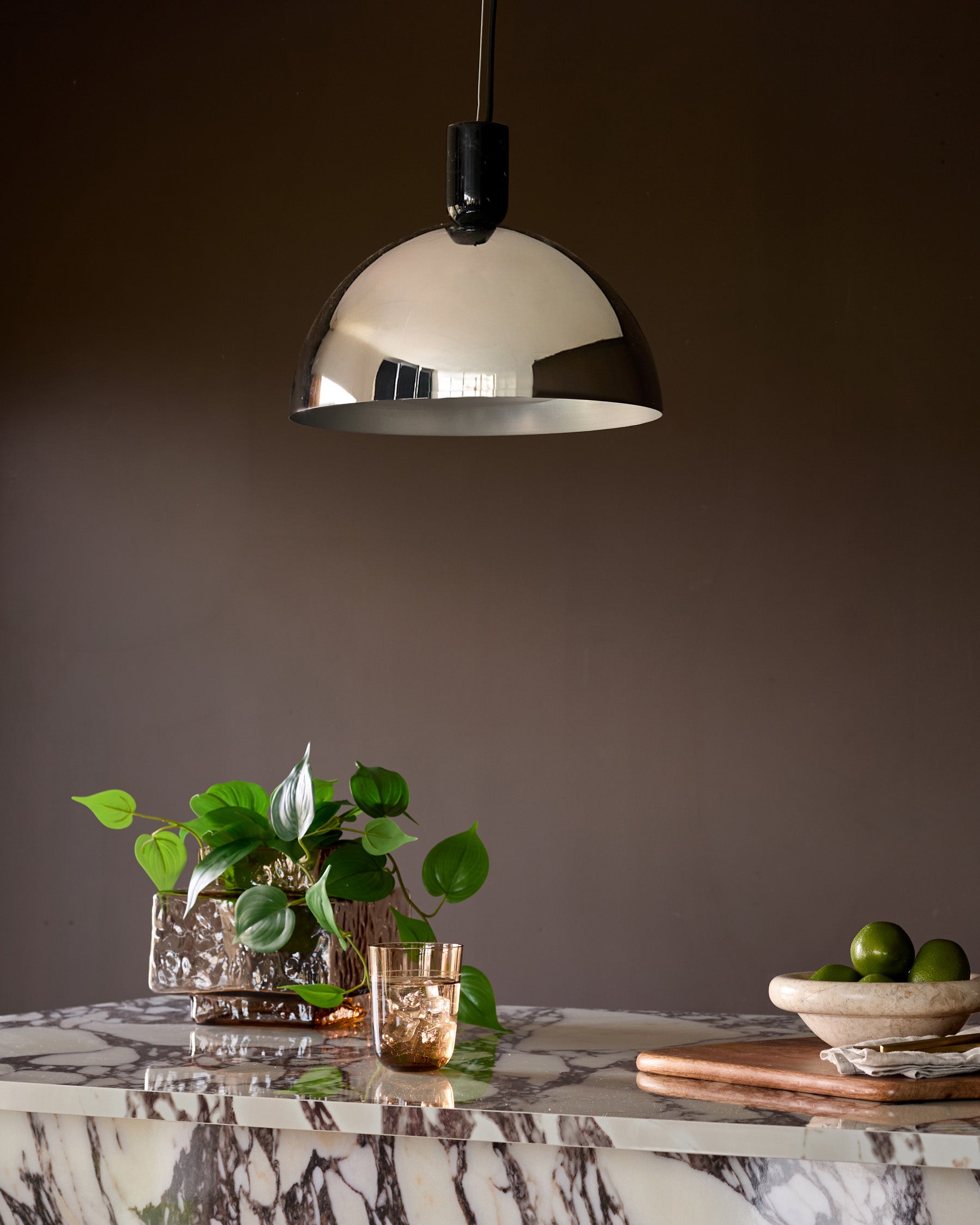 Chrome dome pendant light hanging above marble countertop with plants and fruit bowl.