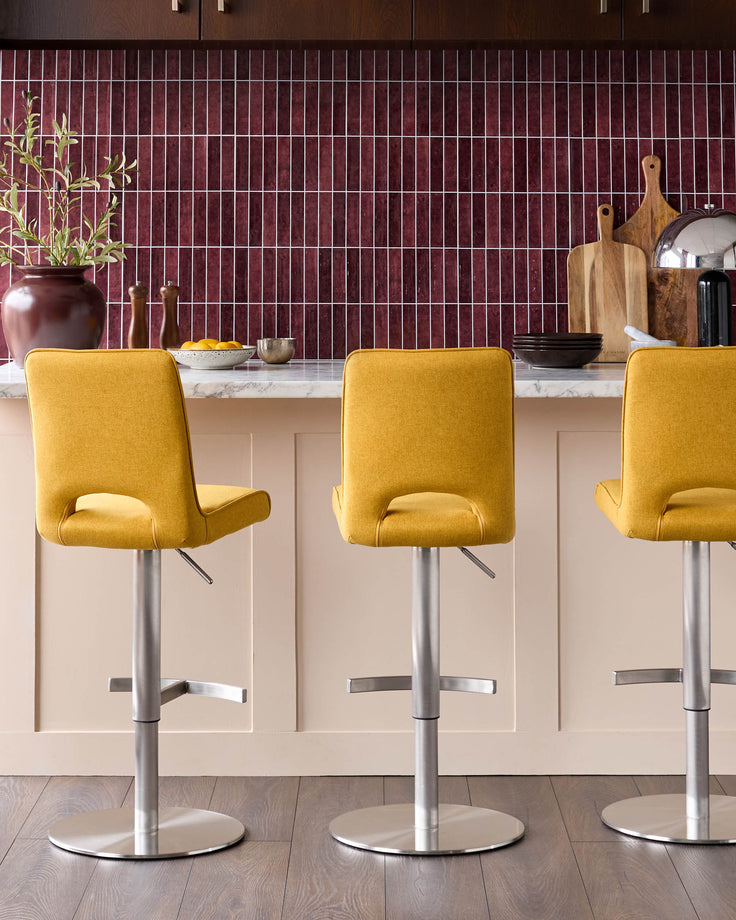 Mustard yellow fabric bar stools with silver gas-lift bases and footrests, shown at kitchen island with burgundy tile backsplash.