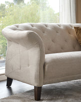 A light beige, button-backed sofa with curved armrests sits in front of a bright window, exuding comfort and style.