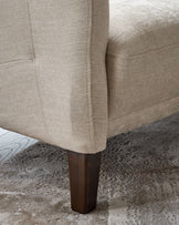 Close-up of a beige armchair with a wooden leg on a patterned rug. Focus on the chair's texture and leg design.