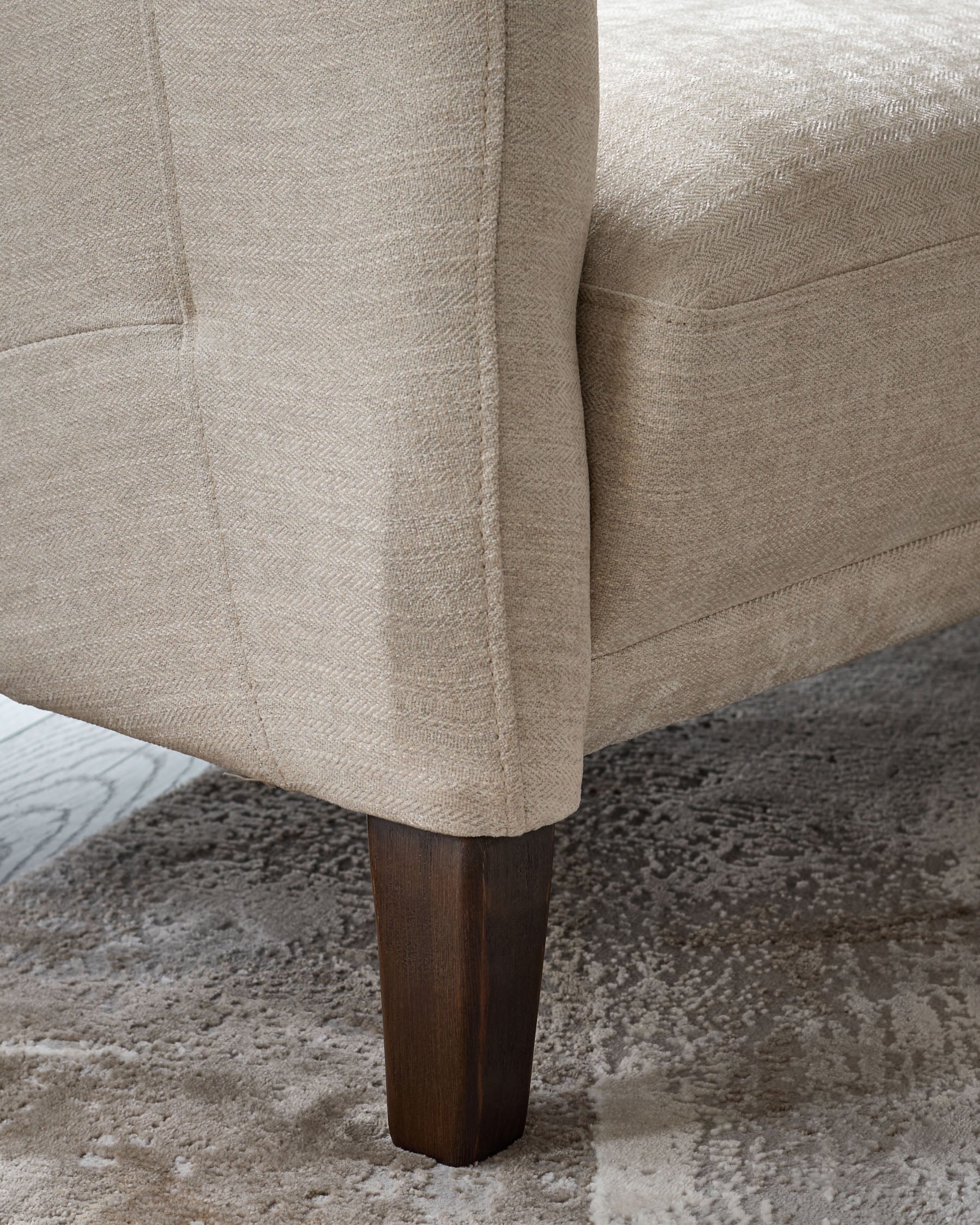 Close-up of a beige armchair with a wooden leg on a patterned rug. Focus on the chair's texture and leg design.