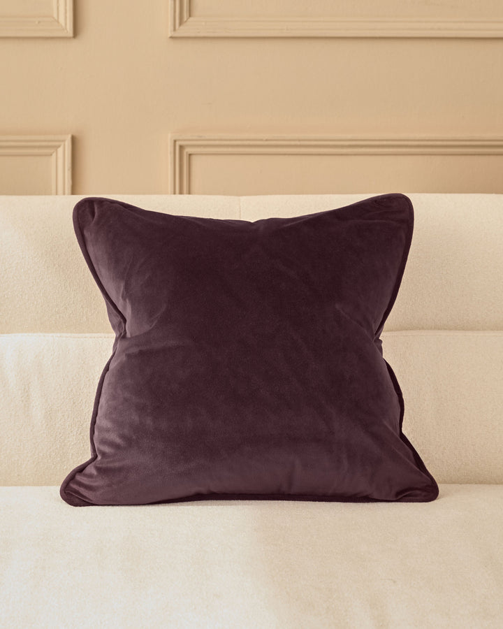 Luca cushion in mulberry velvet, square shape with piped edges, displayed on a cream sofa against a paneled wall.