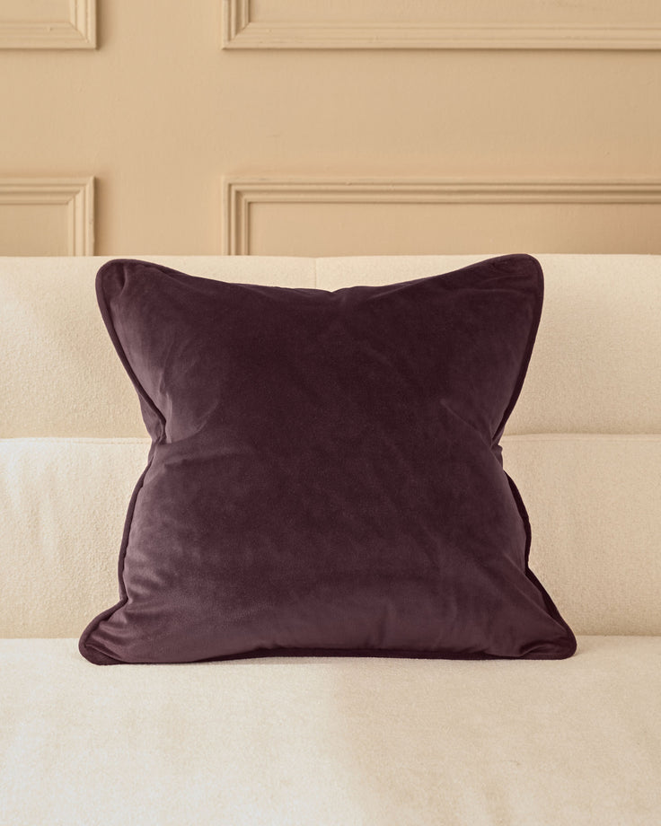 Luca cushion in mulberry velvet, square shape with piped edges, displayed on a cream sofa against a paneled wall.