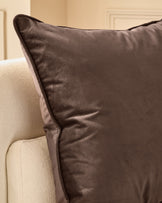 Luca Cushion Medium in chocolate brown, displayed on a cream armchair showing its soft velvet texture and piped edges.