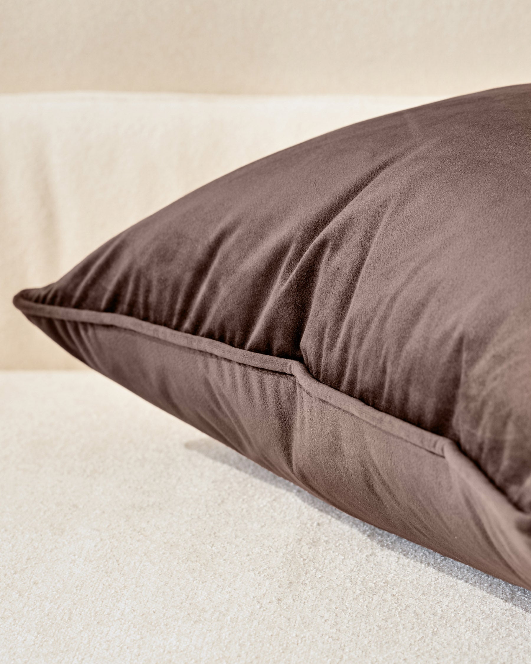 Luca Rectangle Cushion in chocolate brown, shown at an angle on a cream-colored surface, displaying its plump fill and seamed detail.