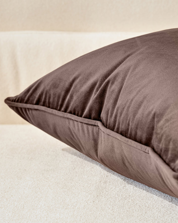 Luca Rectangle Cushion in chocolate brown, shown at an angle on a cream-colored surface, displaying its plump fill and seamed detail.