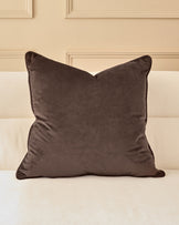 Large chocolate velvet cushion with piping detail, displayed on a cream upholstered bed against a beige paneled wall.