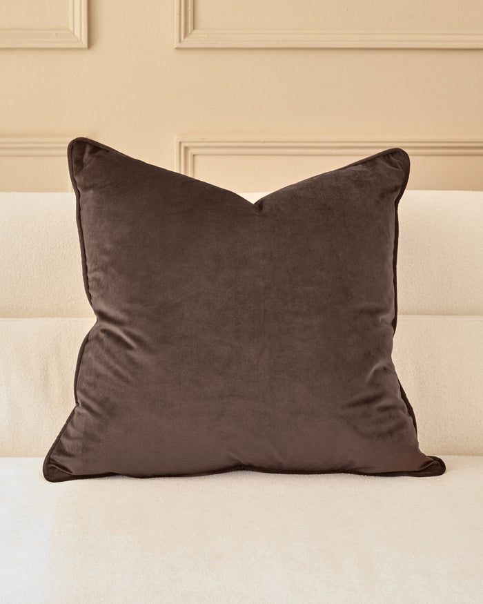 Large chocolate velvet cushion with piping detail, displayed on a cream upholstered bed against a beige paneled wall.